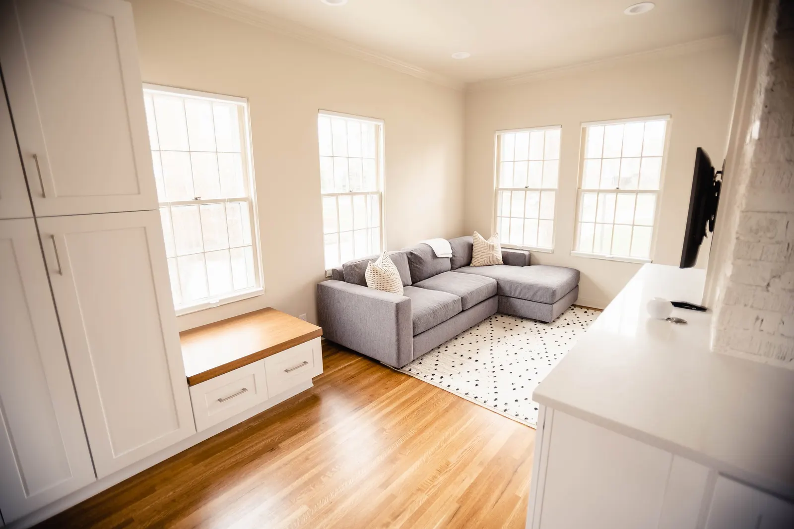 Bright living room with cream walls, hardwood floors, a gray sectional sofa with pillows, three large windows, a white cabinet with a wooden top, and a white rug with black dots.