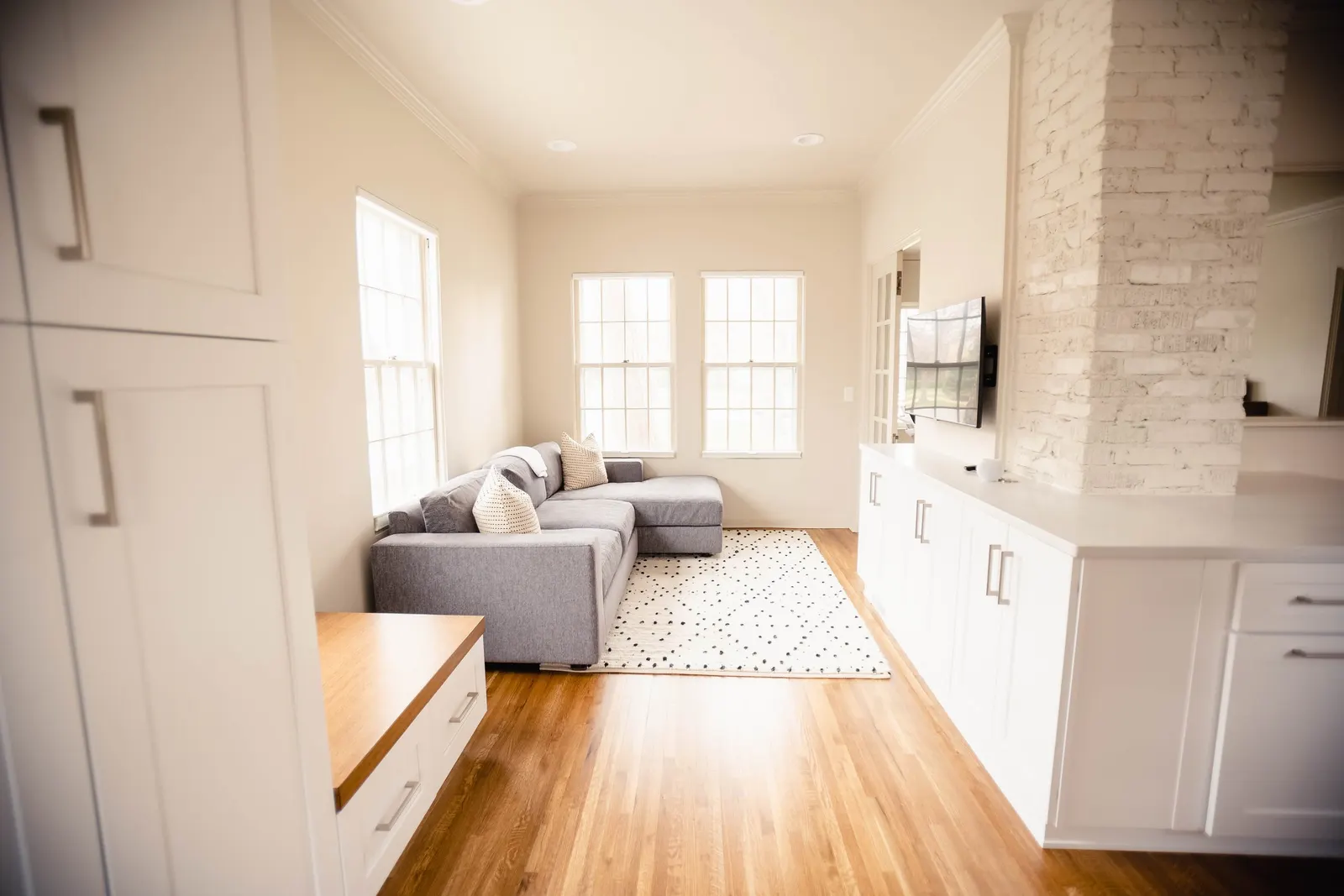 Living room with gray sectional sofa, white cabinets, wooden floor, and three windows with natural light.