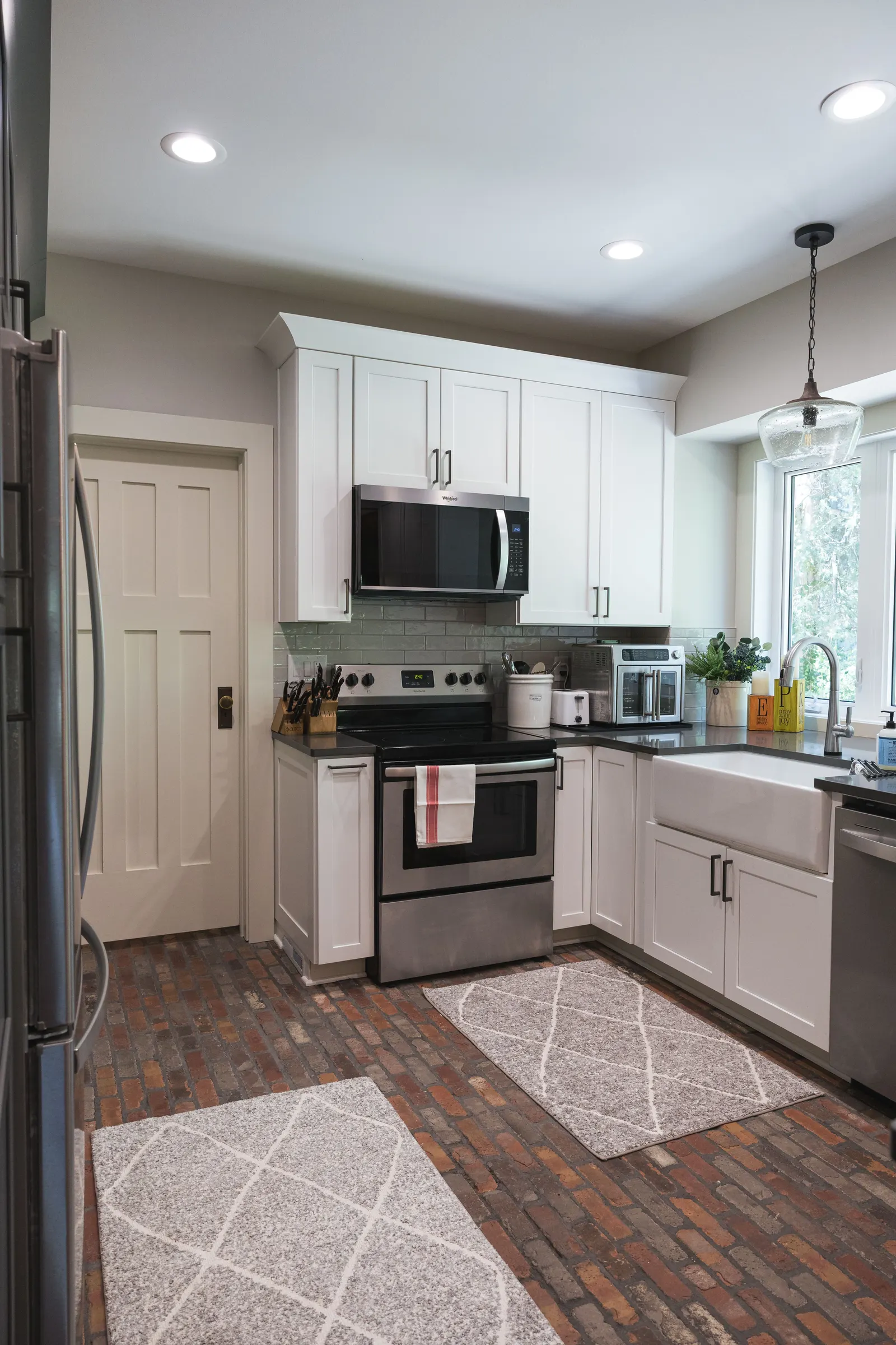 The kitchen from the dining side, looking toward the range wall and the bay window, with a rug on the brick floor.