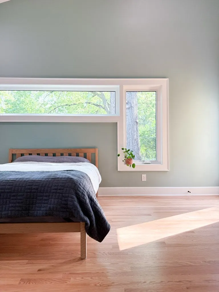 A minimalistic bedroom with a wooden bed frame, a dark quilted blanket, and a large window letting in natural light, with a potted plant on the windowsill and a view of trees outside.