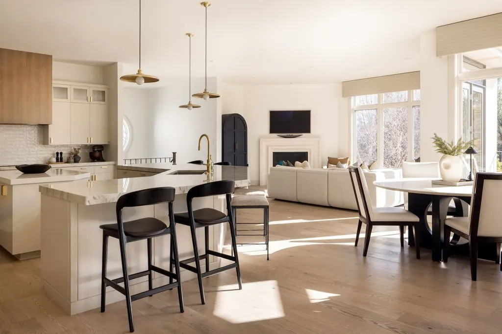 Open-concept living room and kitchen with natural light, white walls, light wood flooring, kitchen island with black bar stools, white cabinetry, dining table with white chairs, and a cozy seating area with a wall-mounted TV above the fireplace.