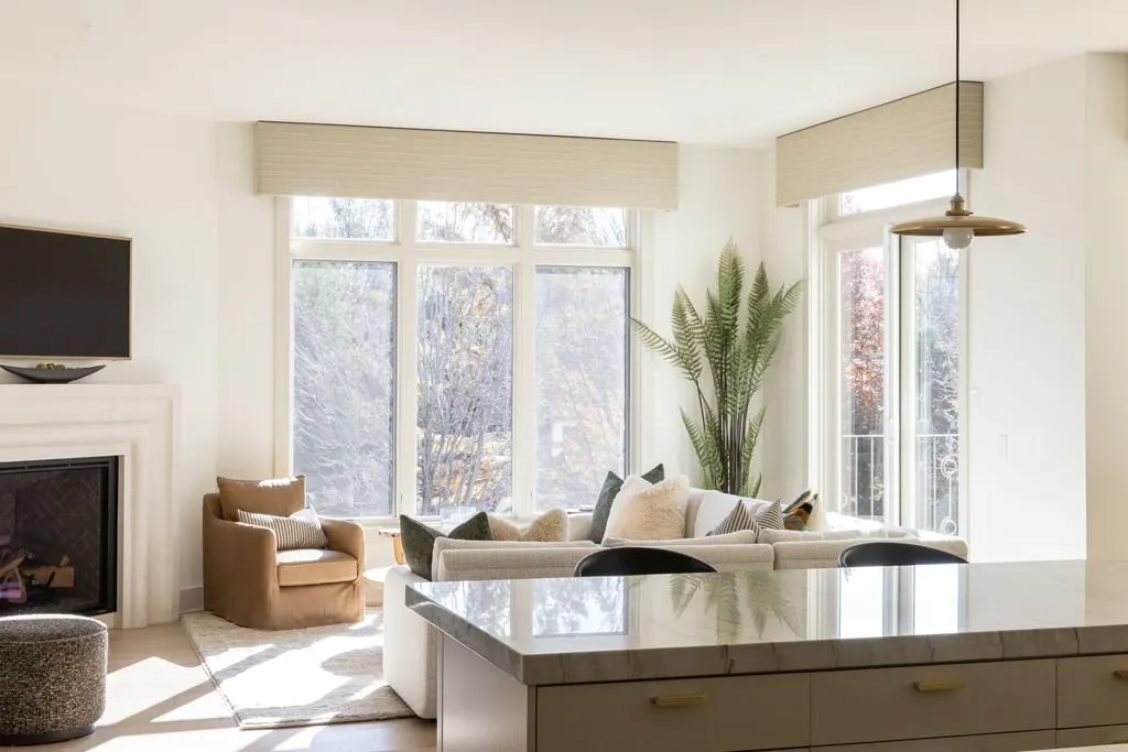 Bright living room with large windows, white sofa, cushioned armchair, fireplace, TV, indoor plant, and kitchen island.