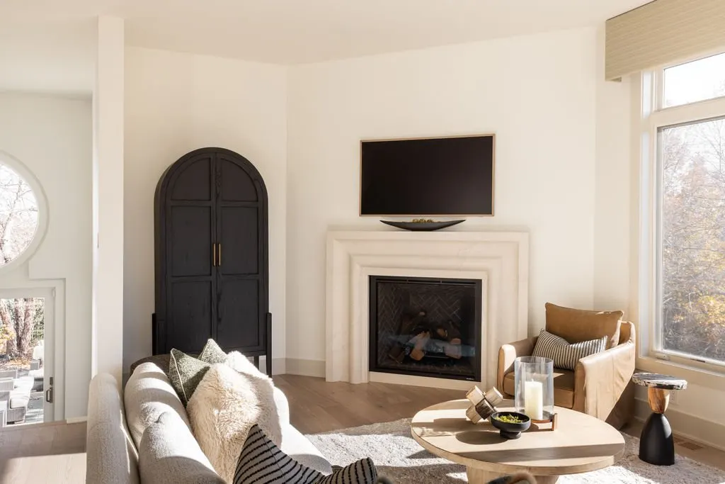 Living room with a beige sofa with pillows, a round coffee table with candles and decor, a beige armchair with pillows, a flat-screen TV above a white fireplace, black cabinet, large window with natural light, and a view of outdoor trees.