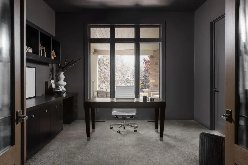 A modern office room with gray walls, a large window overlooking an outdoor balcony, a dark wooden desk with a white chair, a black cabinet with decorative items, and a gray carpet.