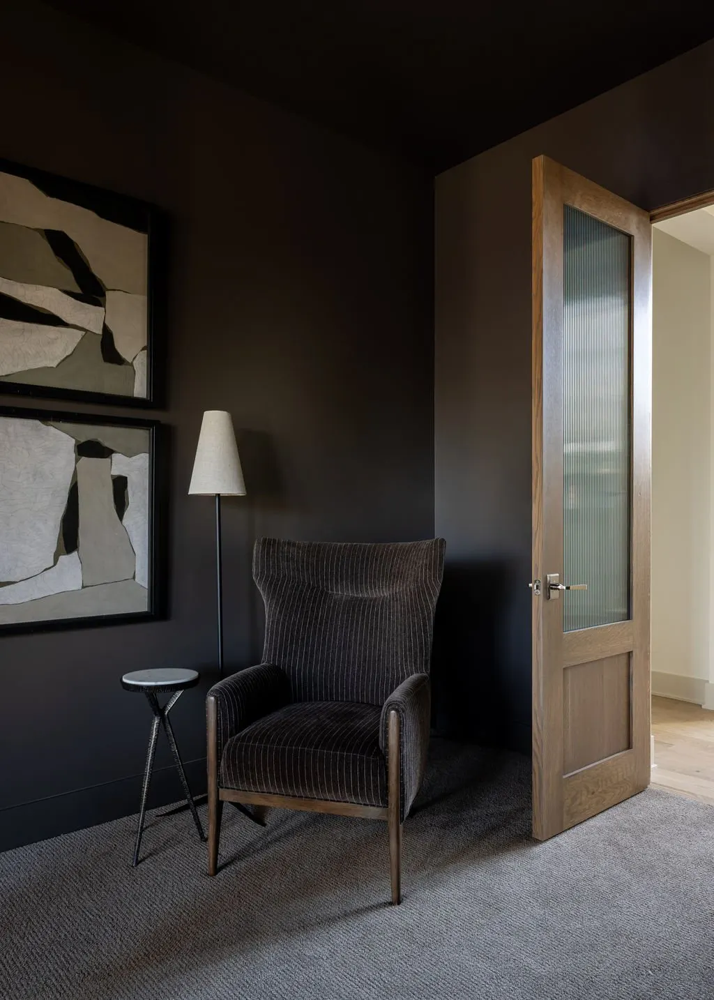 Interior corner of a room with a black wall, a brown upholstered armchair, a side table, a floor lamp, and an open wooden door with glass paneling.