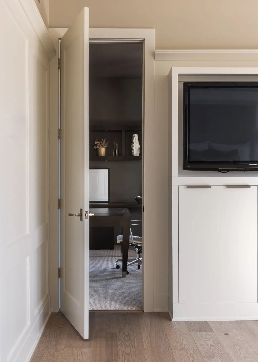 Open door revealing a home office with a desk and chair, partially visible behind a closed white door.