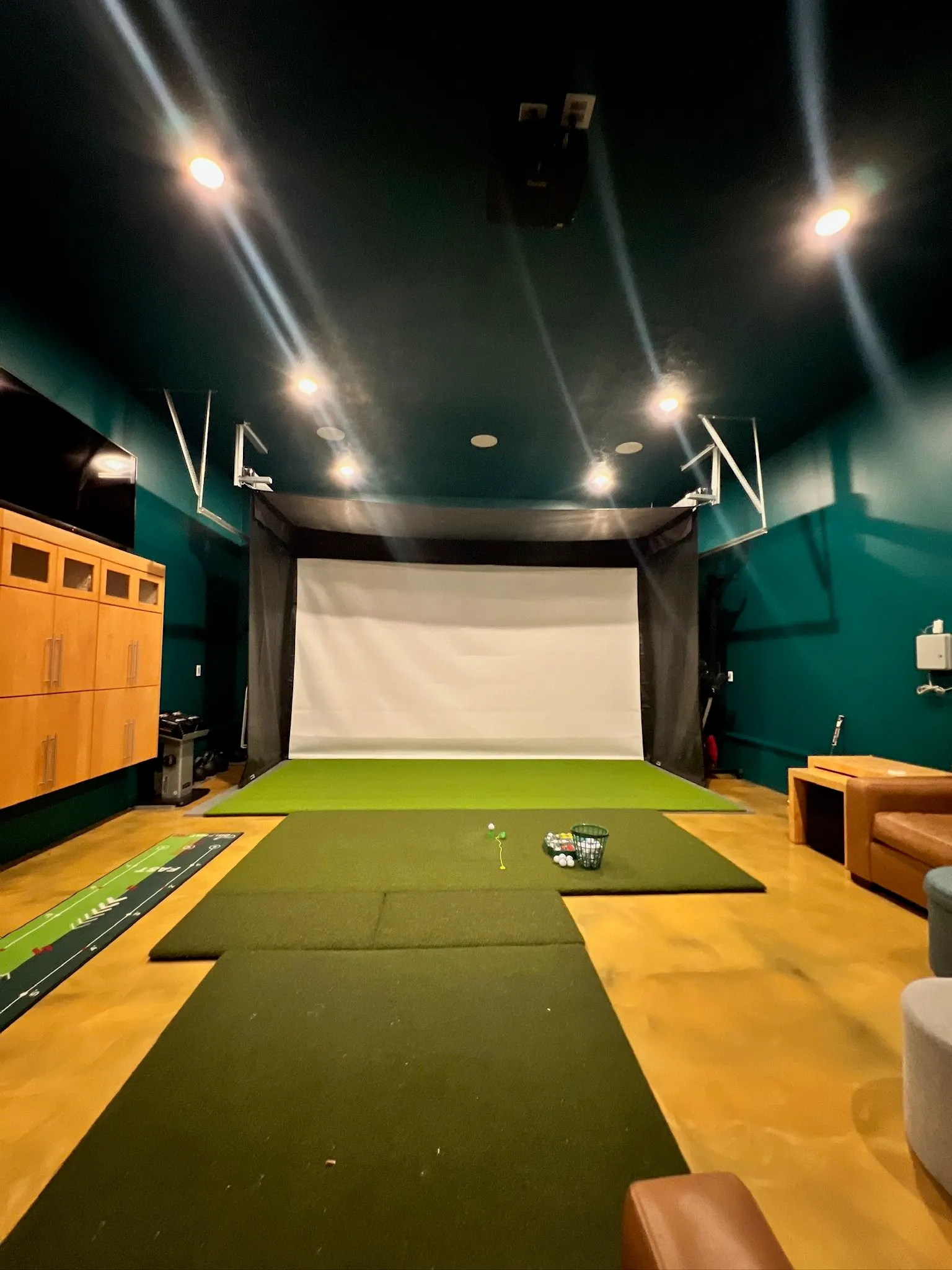 Indoor golf practice room with a large white screen, black walls, and green mats for swing practice. Small golf balls and tees are on the mats, with storage cabinets and a couch visible.