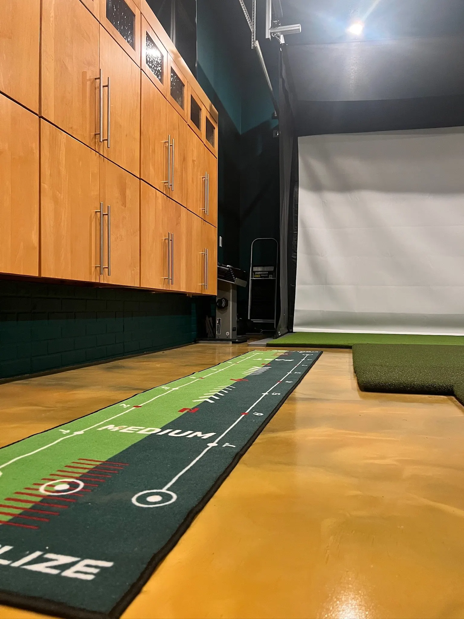 Indoor golf simulator room with a putting green and a radar device on a wooden floor, cabinets on the wall, and a white screen for projection.