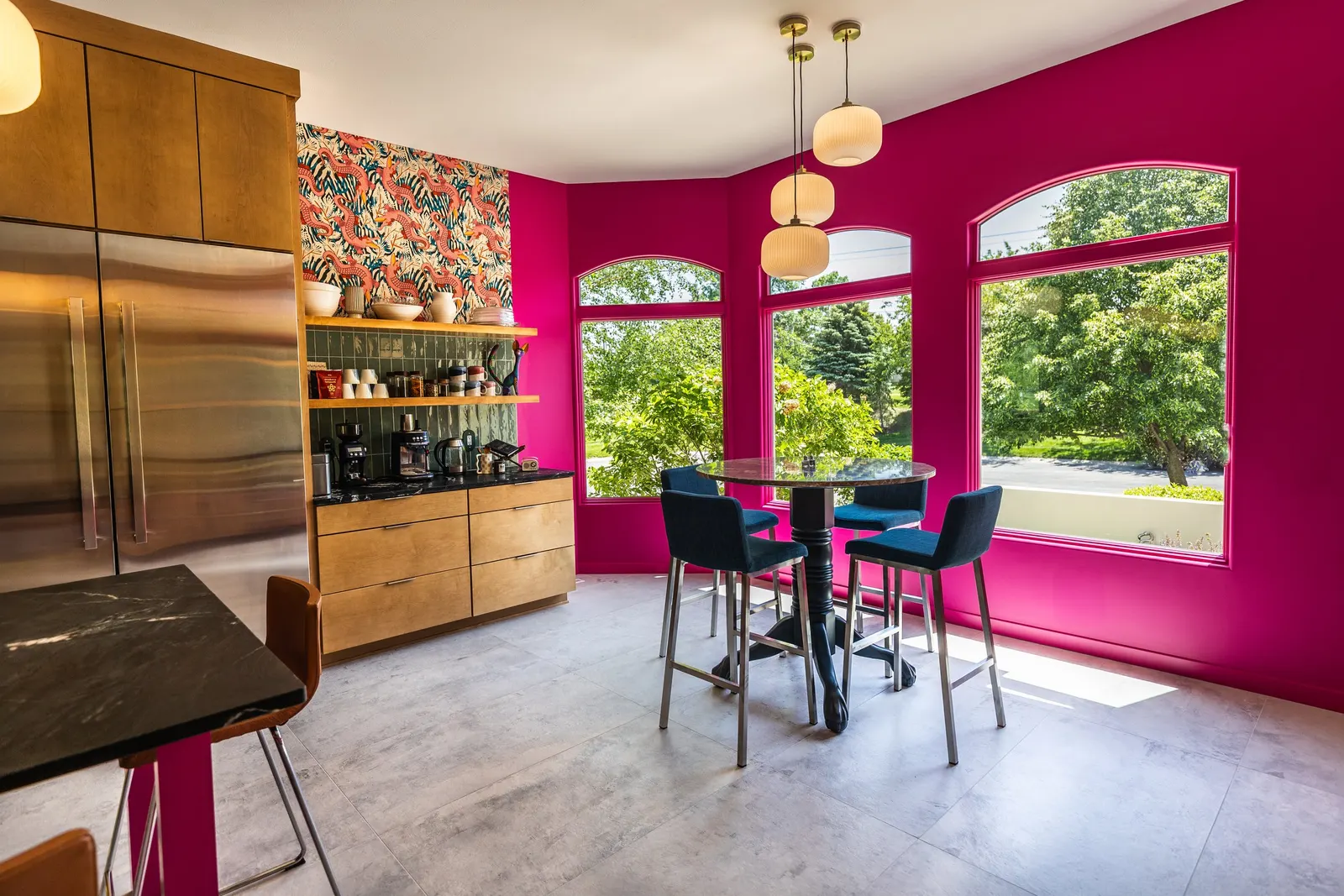 Bright pink kitchen with large windows, black marble table, blue chairs, wooden cabinetry, open shelves with dishes and coffee makers, and colorful flamingo-patterned artwork.