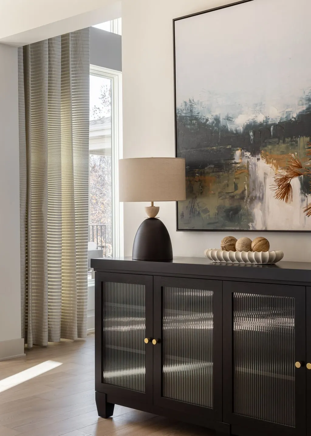 Living room with a black sideboard, a beige table lamp, abstract painting, beige curtains, and a window with trees outside.