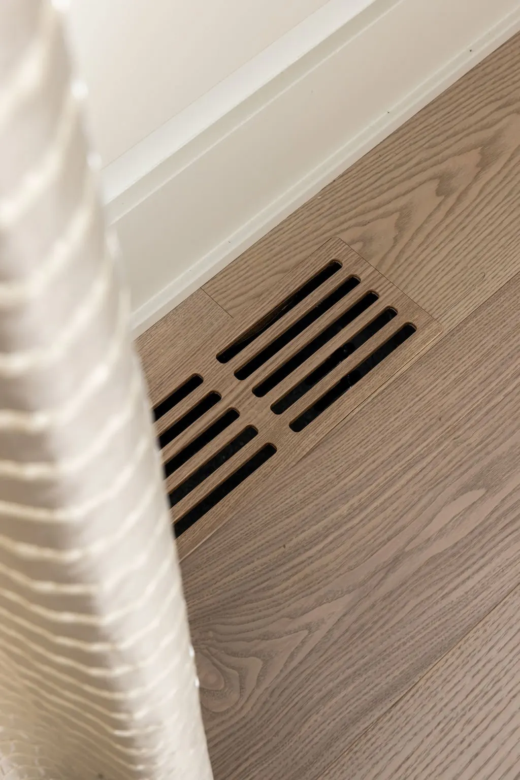 Close-up view of a floor vent with wooden slats, situated on a light wood floor near a white wall and a white curtain.
