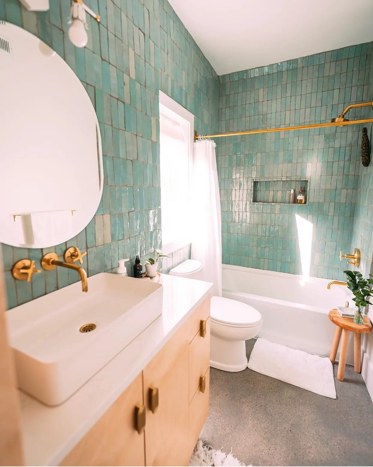 A bathroom with green tiled walls, a white vanity with a ceramic sink, or with brass fixtures, a round mirror above the sink, a window with white curtains, a white bathtub with brass fixtures, a small wooden stool with green plants, and a white bath