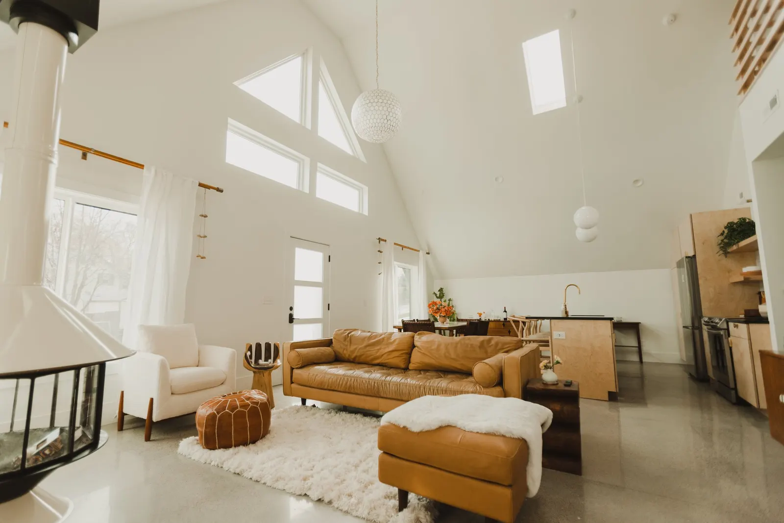 Living room with white walls, high ceiling, multiple windows, and modern furniture including a tan leather sofa, white armchair, orange pouf, and cream shag rug. Kitchen area with light wood cabinets, stainless steel appliances, and a dining table with chairs in the background.