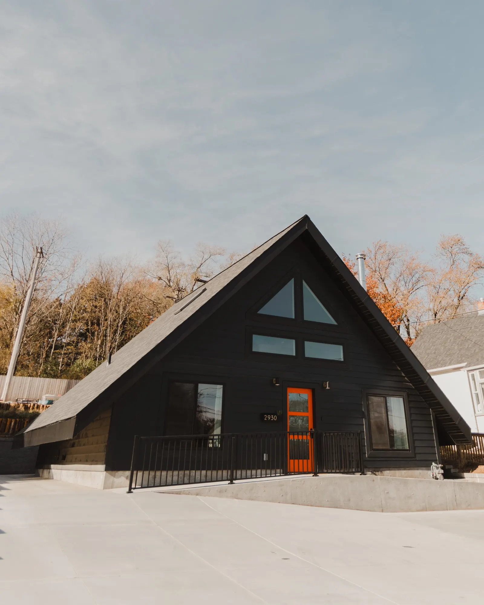 Modern house with black exterior, orange front door, large windows, and a sloped roof, situated on a concrete driveway with a metal railing and surrounded by trees with fall foliage.