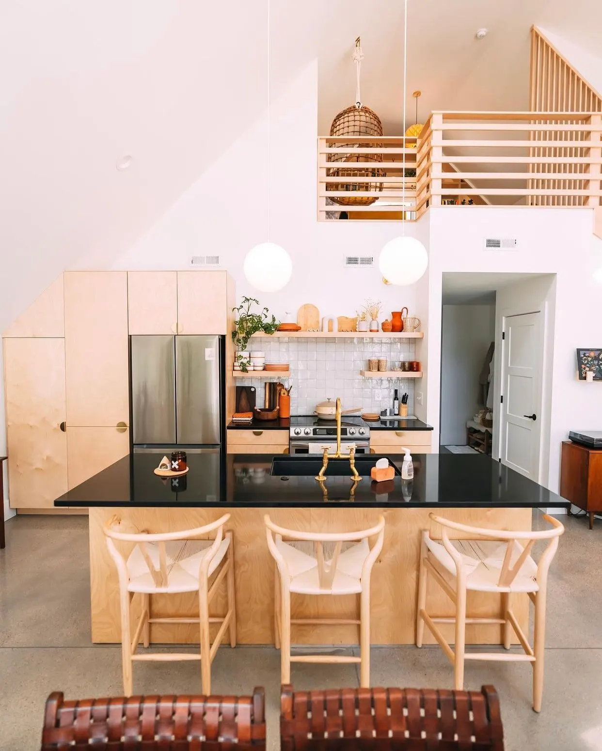 Modern open-concept kitchen with light wood cabinets, black countertop island, and three light wooden chairs with woven seats, high ceiling with two round pendant lights, small shelf with plants and decorative items, stainless steel refrigerator, doo