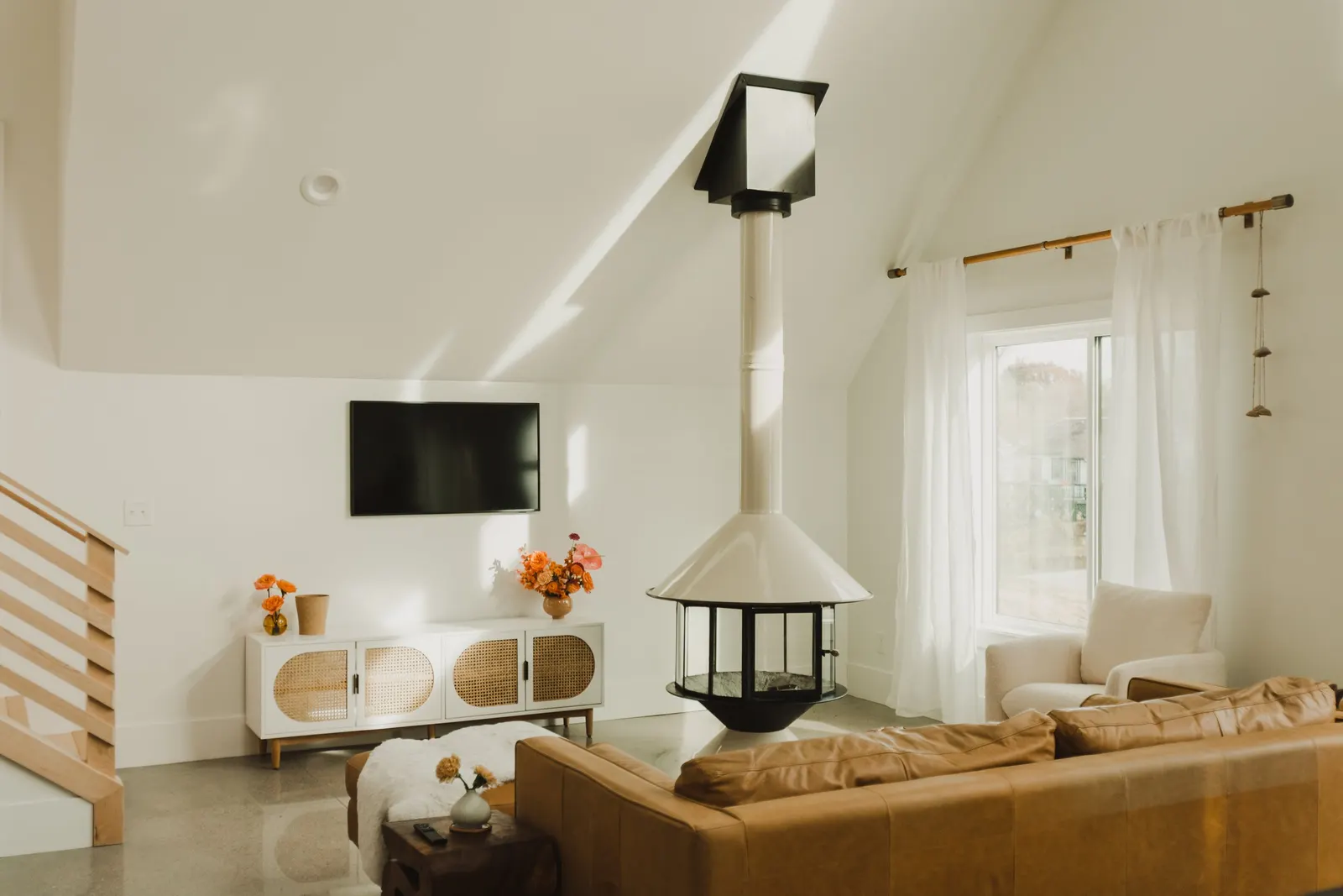 Living room with a beige leather couch, white armchair, wall-mounted TV, a white cabinet with rattan doors, orange and pink flowers in vases, a window with white curtains, a wood curtain rod, and a modern white fireplace with a black chimney.