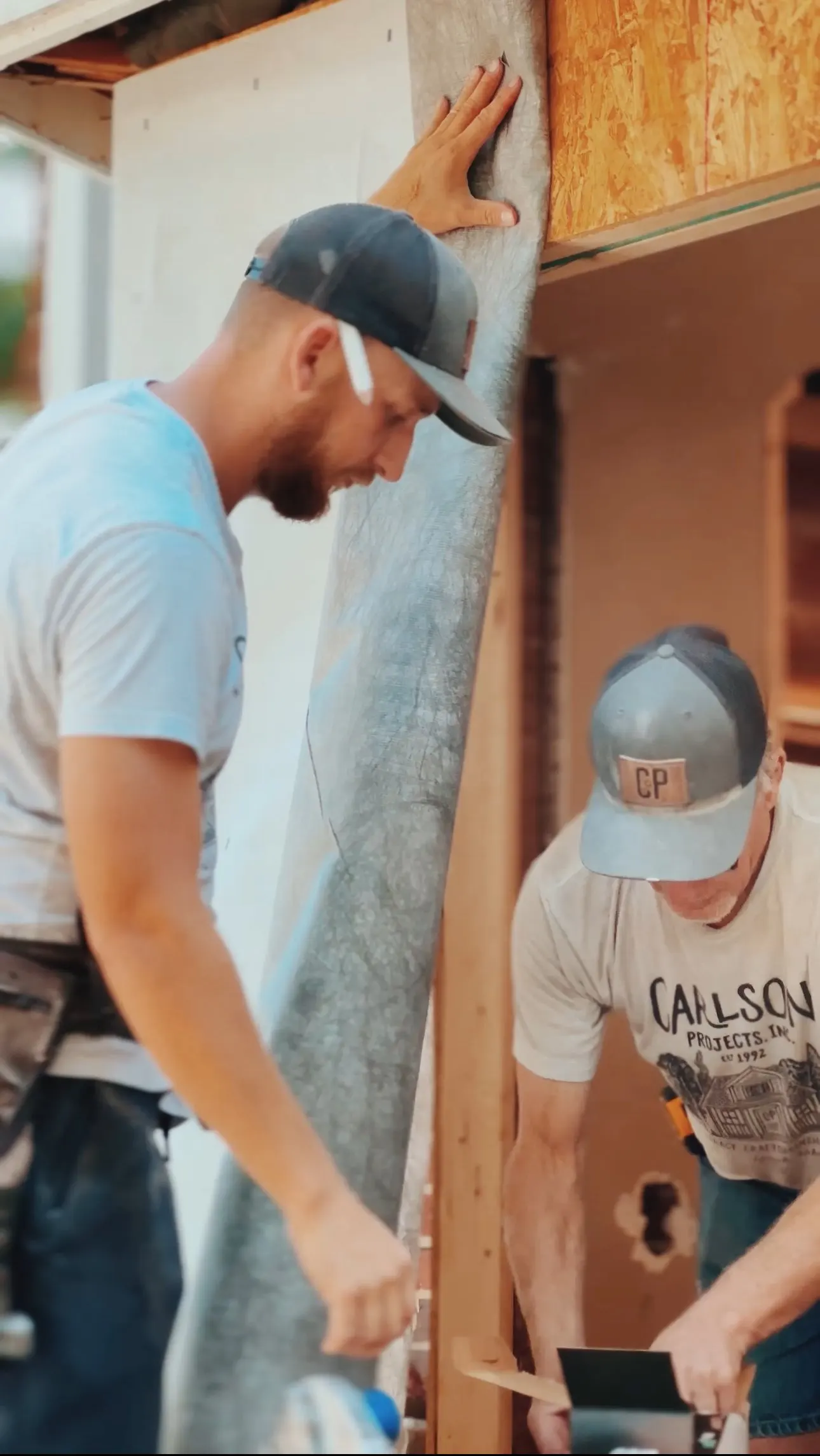 Two construction workers working on a building interior. One is holding a detached section of drywall, and the other appears to be measuring or cutting wood. Both are wearing caps and casual work clothes.