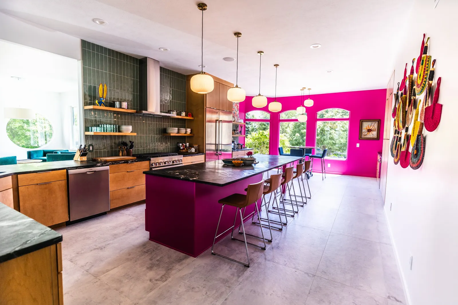 Modern kitchen with a bright pink accent wall, a large island with wooden chairs, green tile backsplash, and colorful hanging art decor.
