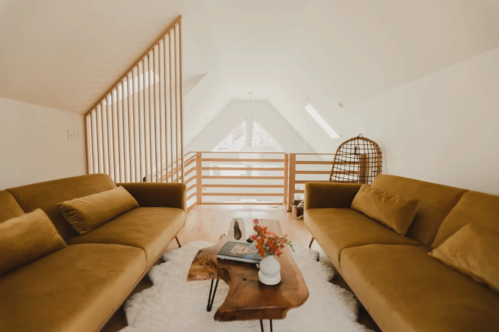 Living room with two brown sofas facing each other, a wooden coffee table with a book and a white vase with red flowers, a white fluffy rug underneath, and a railing overlooking a lower level with a large triangular window.