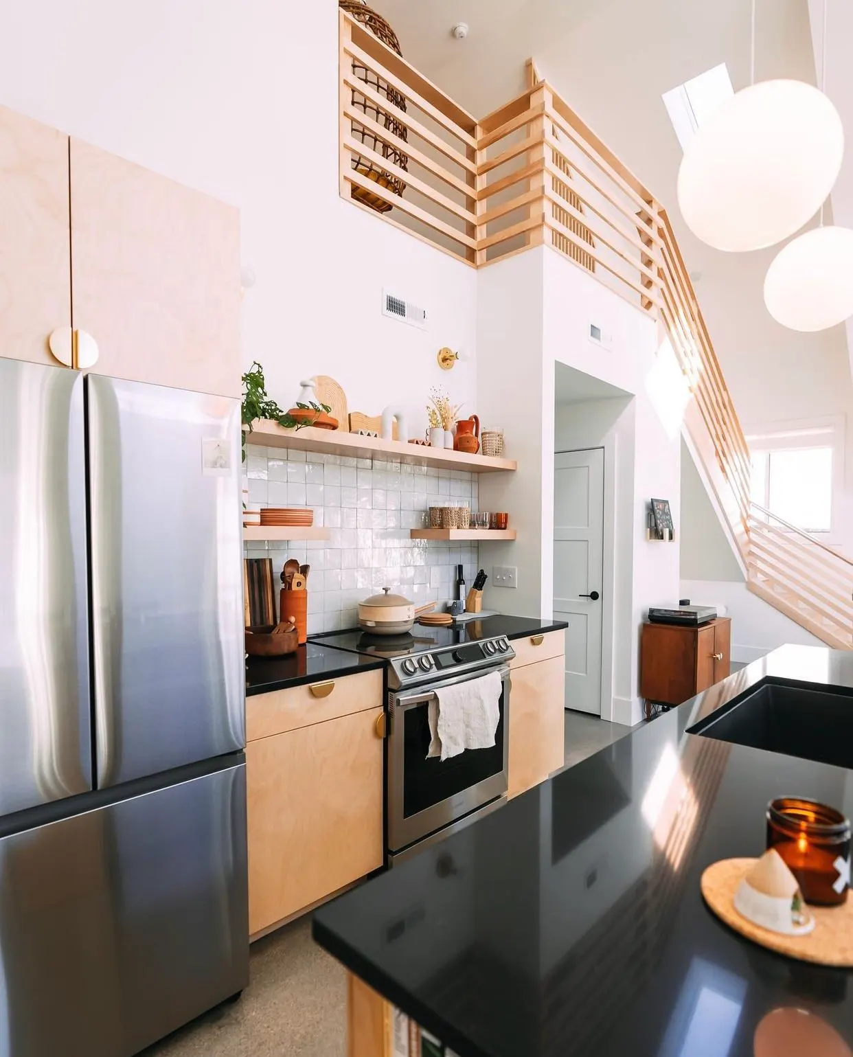 Modern kitchen with black countertops, light wood cabinets, stainless steel refrigerator, stove with a towel, open shelves with decorative items and dishes, white tiled backsplash, and a loft area with wooden railing.