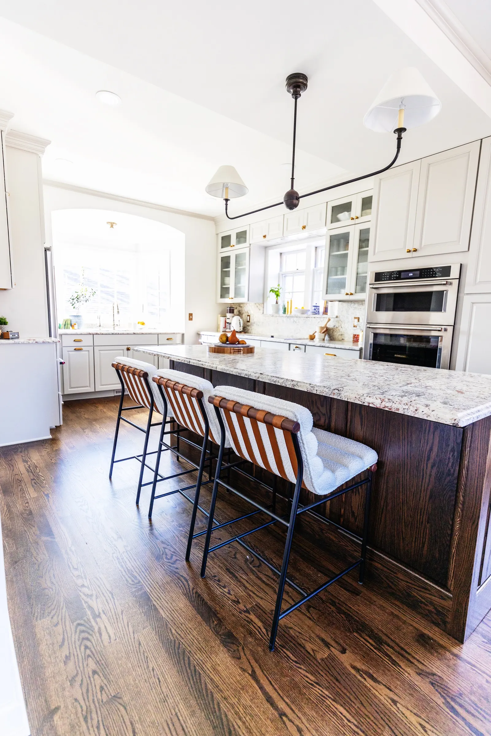 Bright white kitchen with wooden floor, white cabinets, granite countertops, and a kitchen island with bar stools. Windows let in natural light.